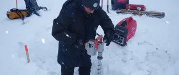 a man drilling a hole in the snow next to a generator