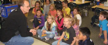 a man kneeling in front of a group of children in a classroom